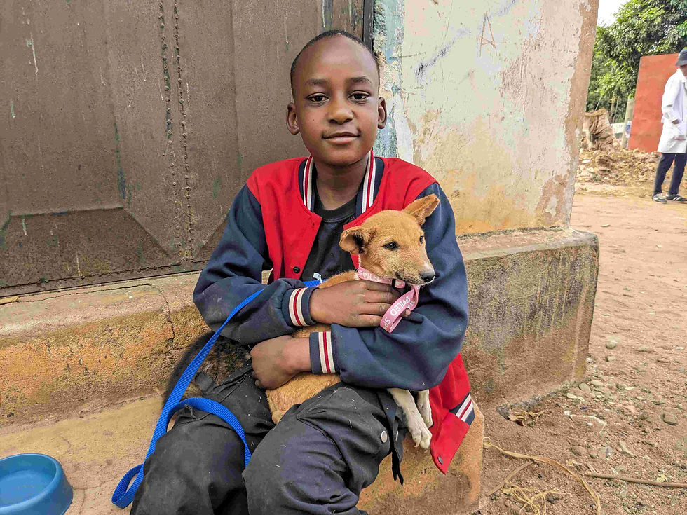 Boy sits outside and holds a small brown dog with a pink collar and blue leash and a bowl is nearby in Tanzania. 