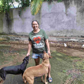 A smiling woman wearing a tshirt that says I proudly save animals stands in a yard with two dogs next to her in Kingston, Jamaica.