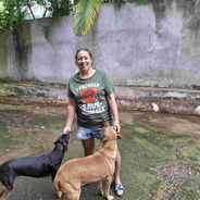 A smiling woman wearing a tshirt that says I proudly save animals stands in a yard with two dogs next to her in Kingston, Jamaica.