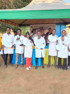 A group of students in white lab coats holding certificats under a green tent in Tanzania.
