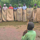 Several women lined up holding burlap sacks and standing on sandy ground with 2 men and a young boy looking at them in Uganda.