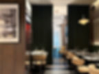 Stylish restaurant interior with beige chairs, black tables, and wine glasses. Dark green curtains, modern chandelier, and wall photos.