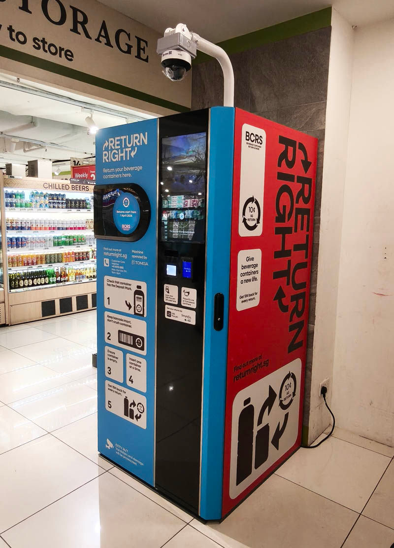 Recycling kiosk in a store with "Return Right" text. Red and blue design, camera above. Shelves of drinks visible in the background.