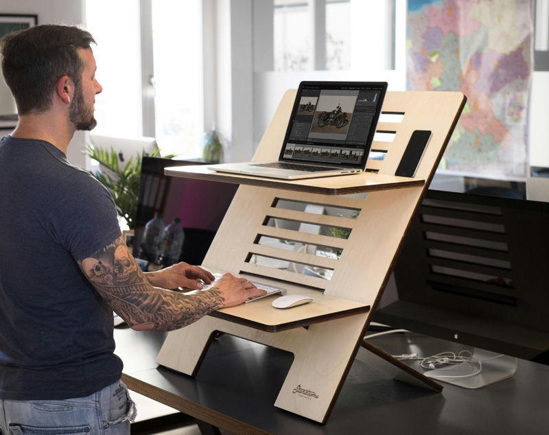 A person uses a laptop on a wooden standing desk in an office. There's a map on the wall and plants nearby, creating a focused atmosphere.