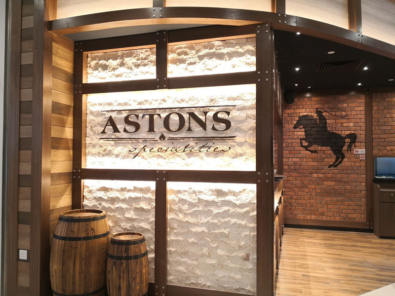 Restaurant entrance with "ASTONS specialties" sign on a lit brick wall. Wooden barrels and horse mural on rustic brick wall in background.