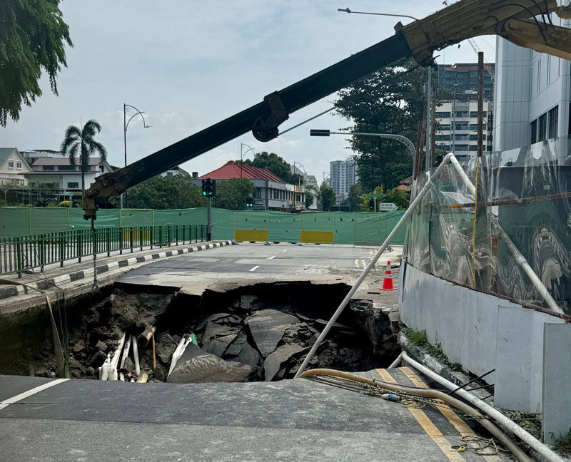 Street with a large sinkhole under repair, surrounded by barriers. A crane is visible. Daytime, with trees and buildings in the background.