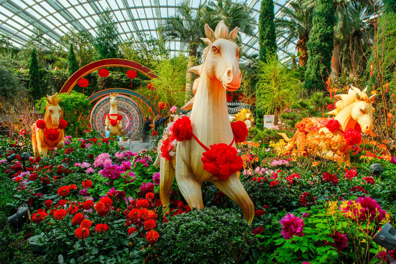 Three wooden horse sculptures adorned with red sashes in a vibrant flower garden with lush greenery under a glass-roofed conservatory.