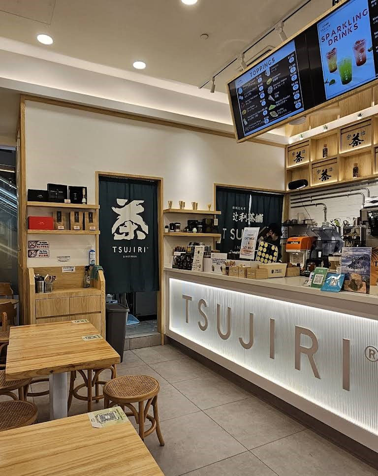 Empty Tsujiri tea shop interior with wooden tables, glowing counter, menu screens, and green noren curtains.