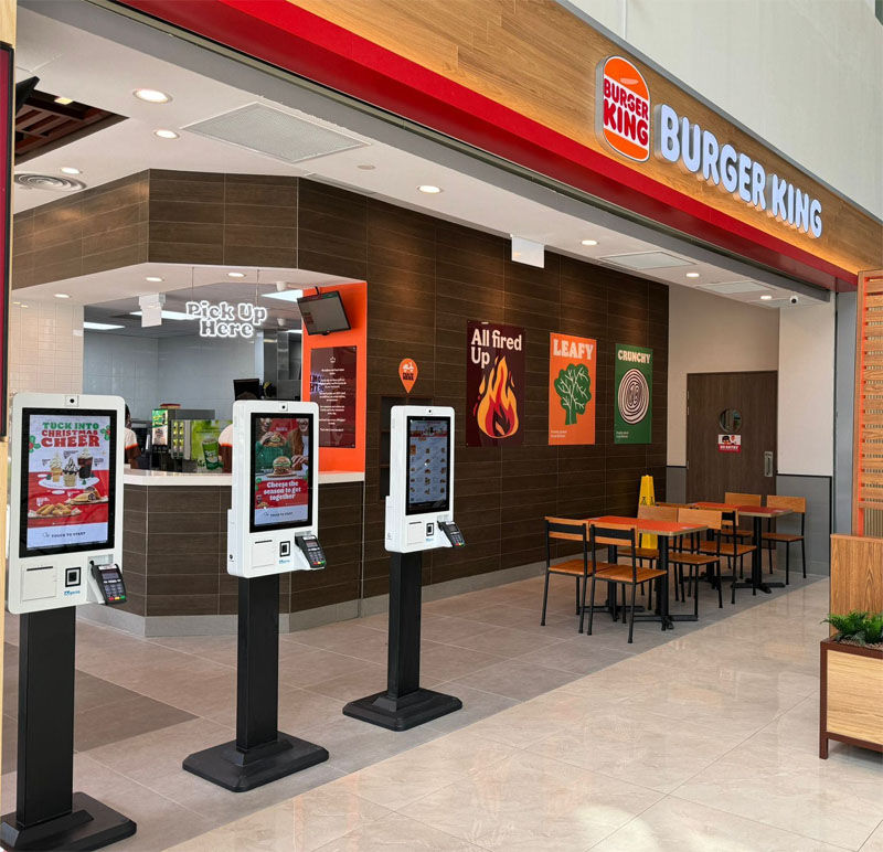 Burger King interior showing three self-service kiosks, a pick-up counter, and posters. Bright lighting, modern decor, and wooden accents.