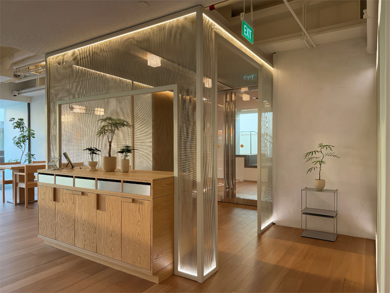 Modern office interior with wooden floors, a glass-paneled meeting room, potted plants, and soft lighting. An "EXIT" sign is visible.