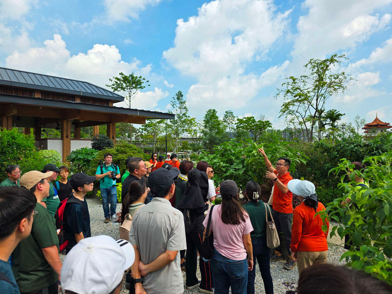 A guide in an orange shirt gestures while leading a diverse group tour in a lush garden. Blue sky and a traditional building in the background.