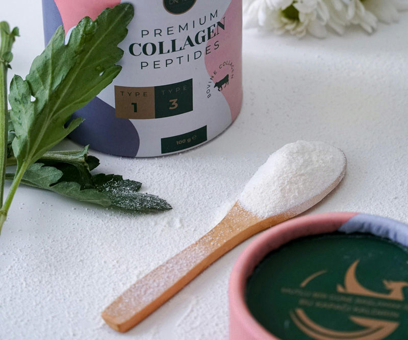 Collagen peptides container, wooden spoon with powder, green leaves, and a white flower on a white surface, creating a fresh and natural vibe.