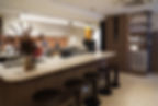 Modern kitchen with a stone island, bar stools, coffee machines, and shelves with jars. Warm lighting and dried flowers create a cozy ambiance.