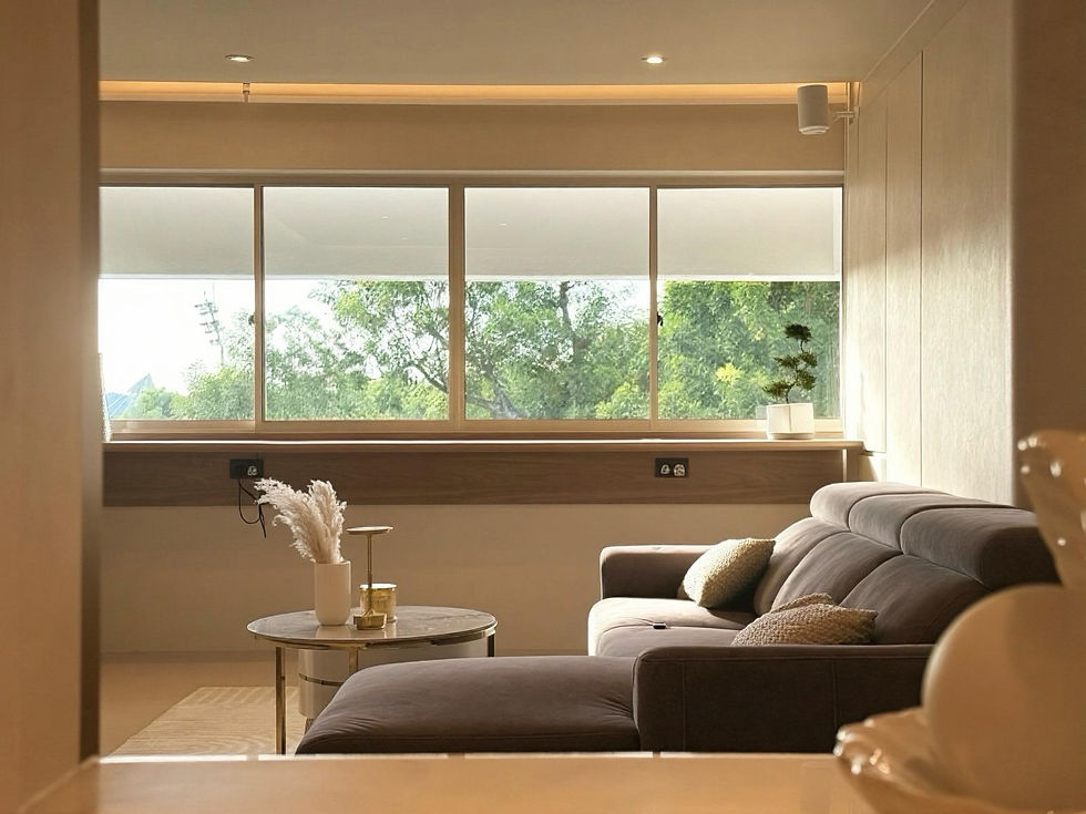 By Wallowin Interior - Cozy living room with a gray sofa, round table with vase and candle, large windows showing trees outside, and soft lighting. Calm atmosphere.