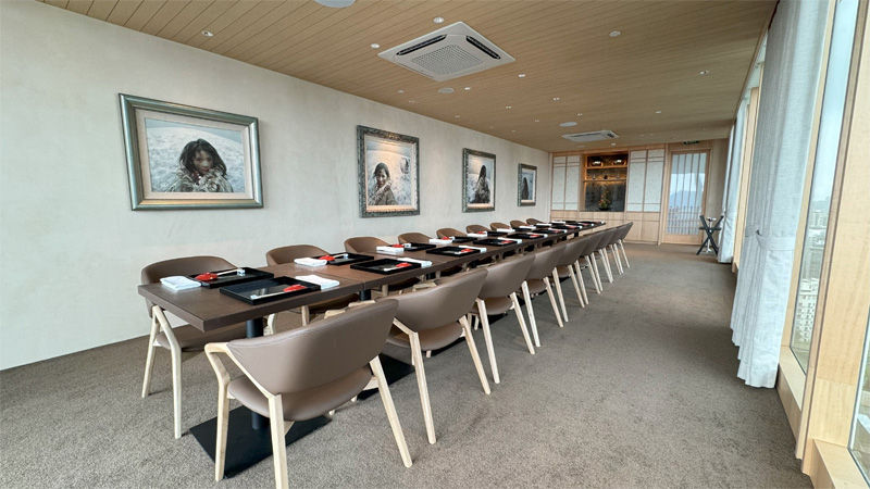 Long conference table set for a meeting in a modern room with beige walls, framed paintings, and large windows. Cozy and professional atmosphere.