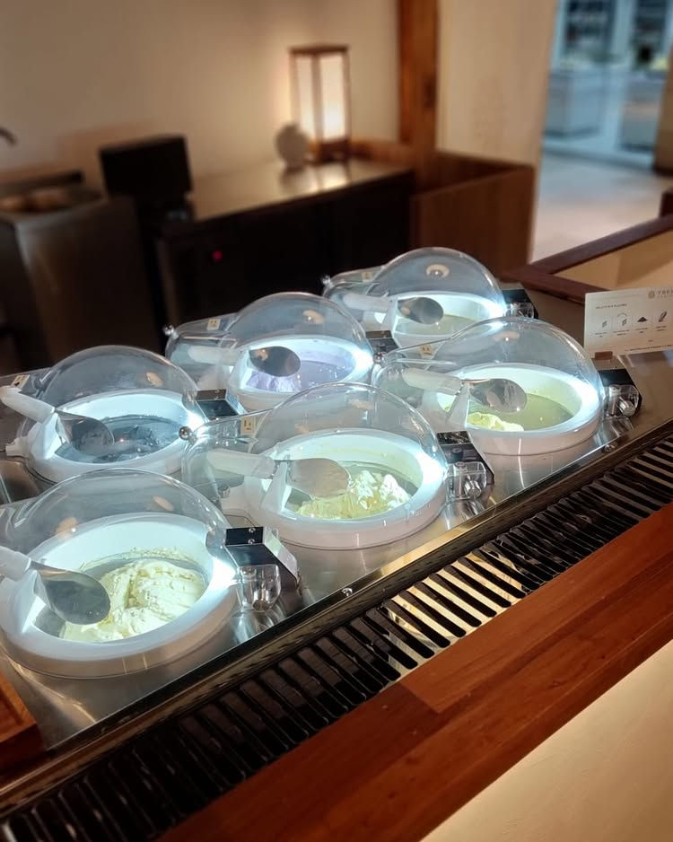 Restaurant buffet with six round, lidded ice cream tubs, scoops inside. Warm lighting, wooden counter. Calm, inviting atmosphere.