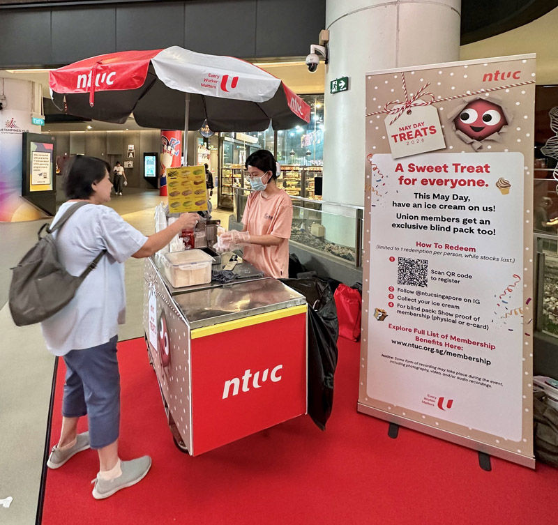 Person at NTUC ice cream stand gives a treat to a customer in a mall. Large sign promotes May Day treats. Red and white theme.