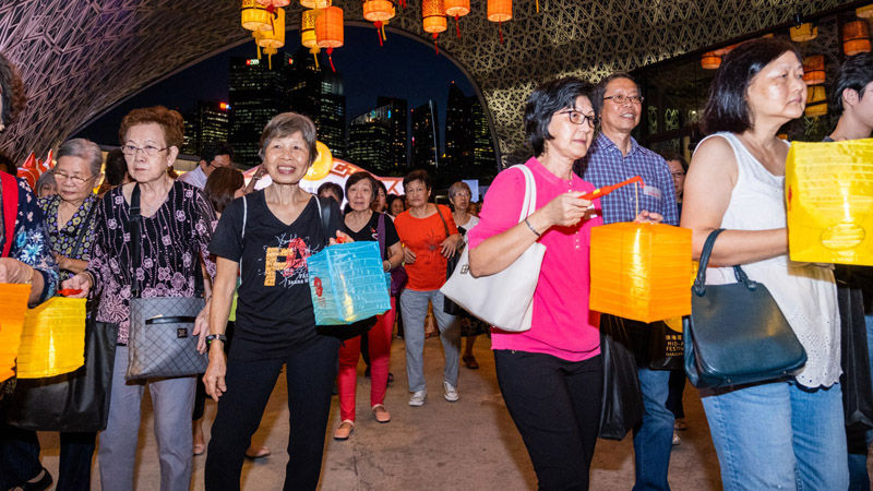 People holding colorful lanterns walk under an illuminated arch at night. Skyscrapers and festive lanterns create a lively atmosphere.
