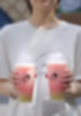 Person holding two pink and green gradient drinks in clear cups with a logo, wearing a white t-shirt, sunny outdoor setting.