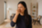 A woman pinches her nose, appearing displeased, in a cozy living room with neutral tones, a sofa, lamp, and shelf in the background.