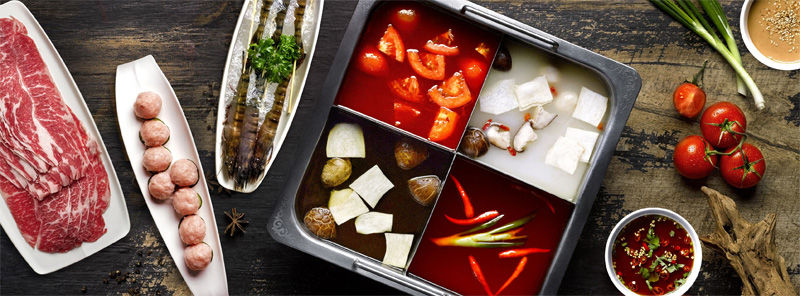 A divided hot pot with red and clear broths, surrounded by raw meats, shrimp, vegetables, and spices on a rustic wooden table.