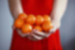Hands hold a bunch of bright orange mandarins against a red dress background, creating a vibrant contrast. Mood: fresh and inviting.
