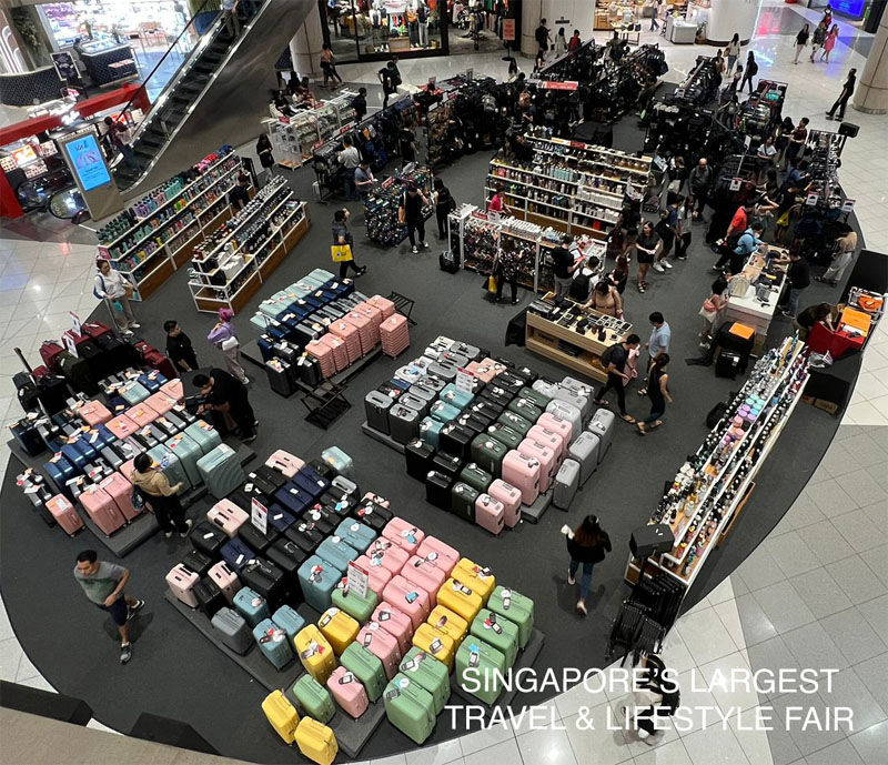 Overhead view of a bustling travel fair with colorful luggage and display stands. Shoppers explore. Text: Singapore's Largest Travel & Lifestyle Fair.
