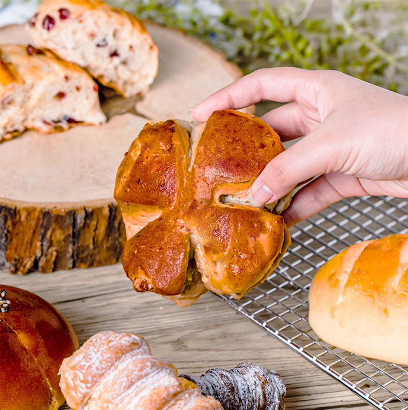 A hand holds a clover-shaped bun over a wooden table with assorted pastries and a wire rack. Background shows greenery.