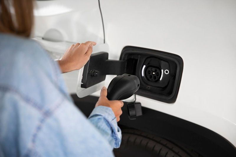 Person in a denim jacket plugging a charger into a white electric car's charging port. The setting is bright and the mood is focused.