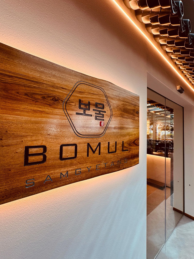 Wooden sign with Korean characters and "BOMUL SAMGYETANG" text on a wall, under warm lighting. Glass door on the right. Calm ambiance.