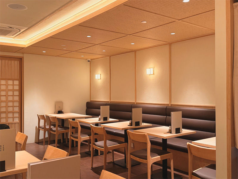 Empty restaurant with wooden tables and chairs, cream walls, and soft overhead lighting. Menus stand upright on the tables. Cozy ambiance.