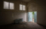 Dim room with tiled walls, barred windows, and open blue double doors leading outside. An old radiator lies on the floor. Quiet and abandoned.