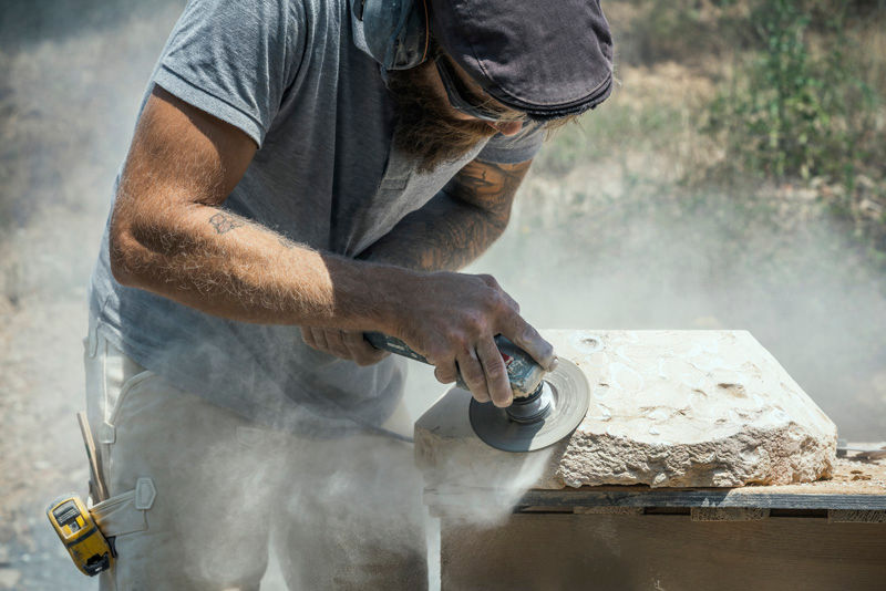 A person wearing a cap and earmuffs uses a power tool to carve a stone slab, surrounded by dust, in an outdoor setting.