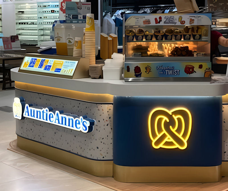 Auntie Anne's kiosk with pretzels in a display, neon pretzel sign, branded cups, terrazzo countertop, and menu screen in a shopping mall.