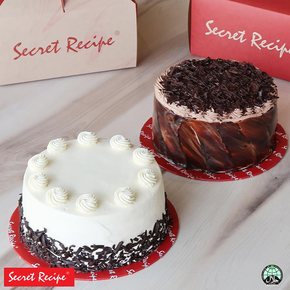 Two cakes on a table: a white cake with chocolate shavings and a chocolate cake with chocolate flakes. Boxes labeled "Secret Recipe" in the background.