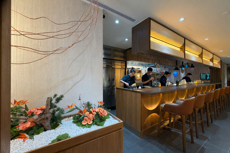 Chefs work at a wooden sushi bar with orange seating, under warm lighting. A floral arrangement with orange flowers decorates the foreground.