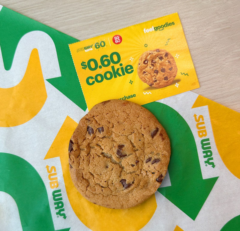 Chocolate chip cookie on a Subway sandwich wrapper. A yellow coupon offers a $0.60 cookie deal. Bright colors, casual setting.