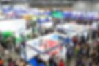 Crowded expo hall with colorful booths and banners. People browse travel exhibits. ClubMed and Changi signs visible. Busy and vibrant scene.