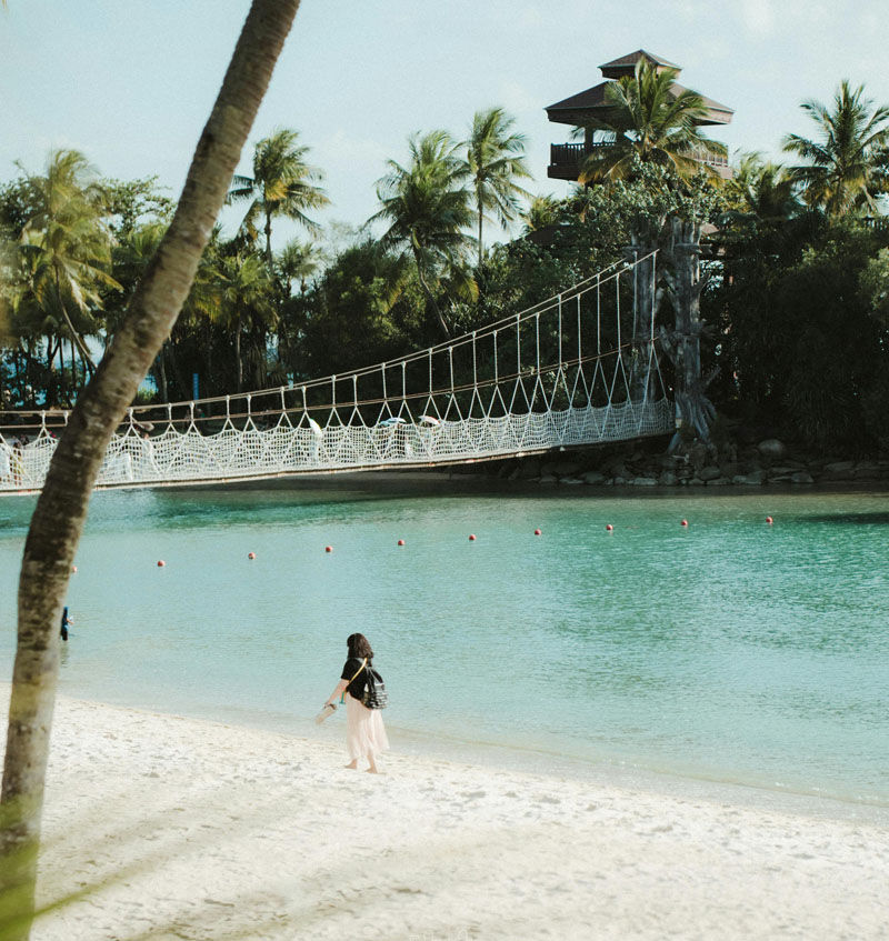 Person walks on a beach by turquoise water and a rope bridge. Palm trees and a wooden structure in the background under a clear sky.