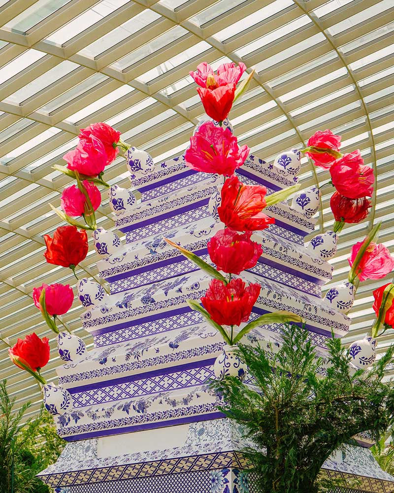 Vibrant red tulips adorn a patterned, blue-and-white structure beneath a glass ceiling. Greenery surrounds the base, creating a lively ambiance.