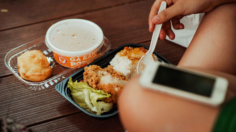 Person eating fried chicken, rice and soup from a takeout tray on a wooden table, with a phone nearby.