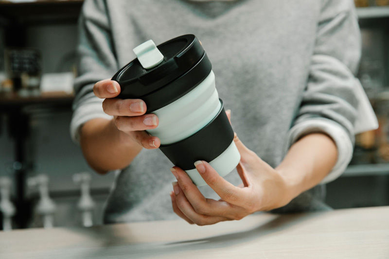 Person in gray sweater holding a black-and-white collapsible travel cup at a counter in a cozy cafe or kitchen.