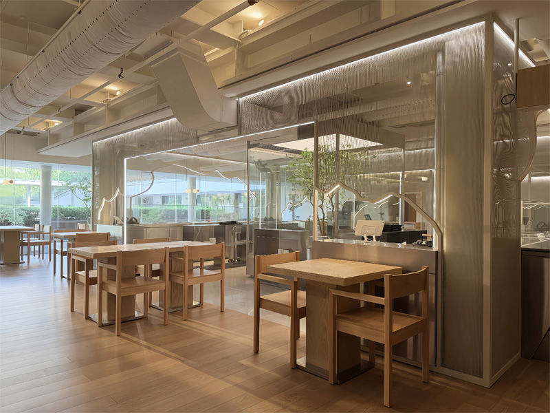 Modern cafe with light wood tables and chairs. Large glass partitions and greenery create an airy feel. Bright natural light streams in.