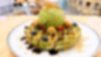 Matcha waffle topped with green ice cream, blueberries, strawberries, nuts, and chocolate sauce on a white plate in a cafe setting.