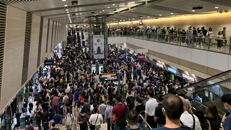 Crowded subway station with people moving on escalators and platforms. Signs display train info. Busy, bustling atmosphere.