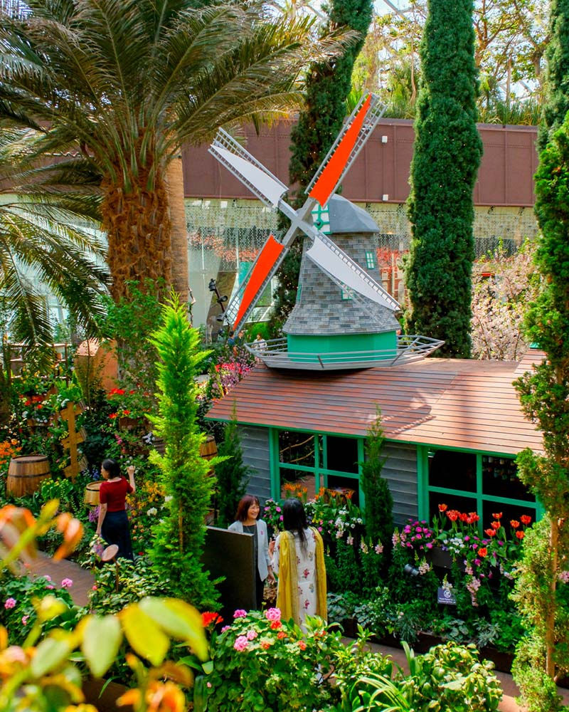 Colorful garden with a windmill and lush greenery. Three people chat among vibrant flowers, creating a serene, lively atmosphere.