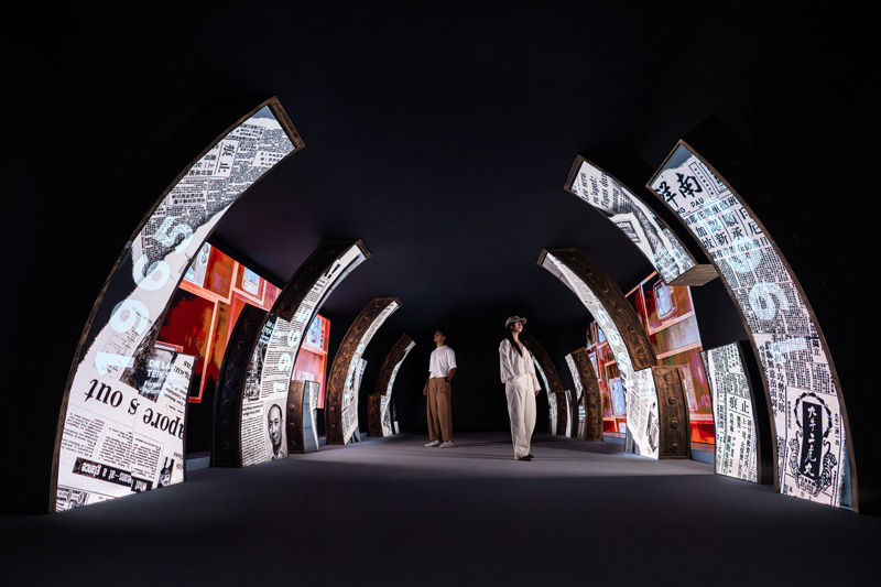 Two people walk in a dark, arched hallway with illuminated panels displaying newspapers and red accents, creating a futuristic feel.