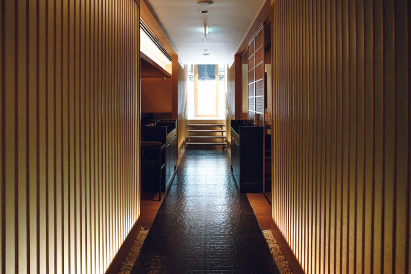 Narrow corridor with wooden walls, dark floor, and stone accents leads to a bright window and stairs. Mood is calm and empty.