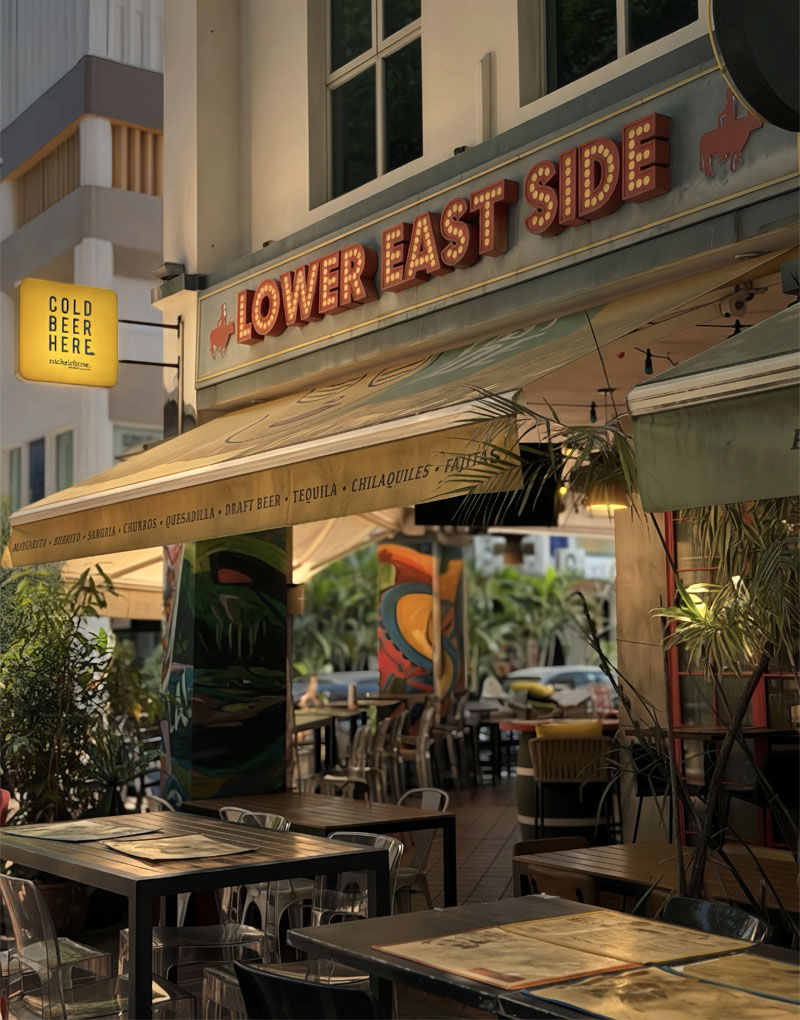 Outdoor café with a "LOWER EAST SIDE" sign, under yellow awning. Text "COLD BEER HERE" is visible. Tables and chairs are empty, warm ambiance.