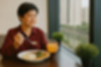 Woman in red shirt eats rice dish with broccoli, holds orange juice. Seated by window with cityscape and plants, calm setting.